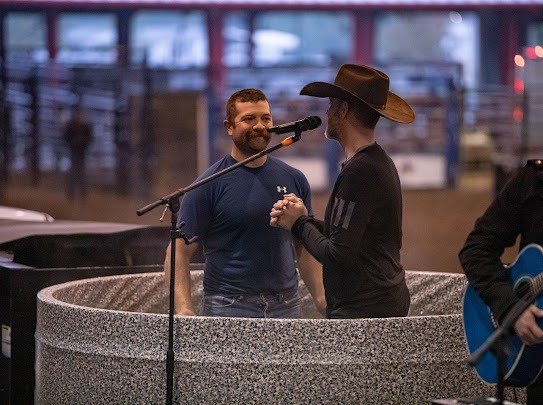 A man being baptized in an arena while a pastor in a cowboy hat shakes his hand