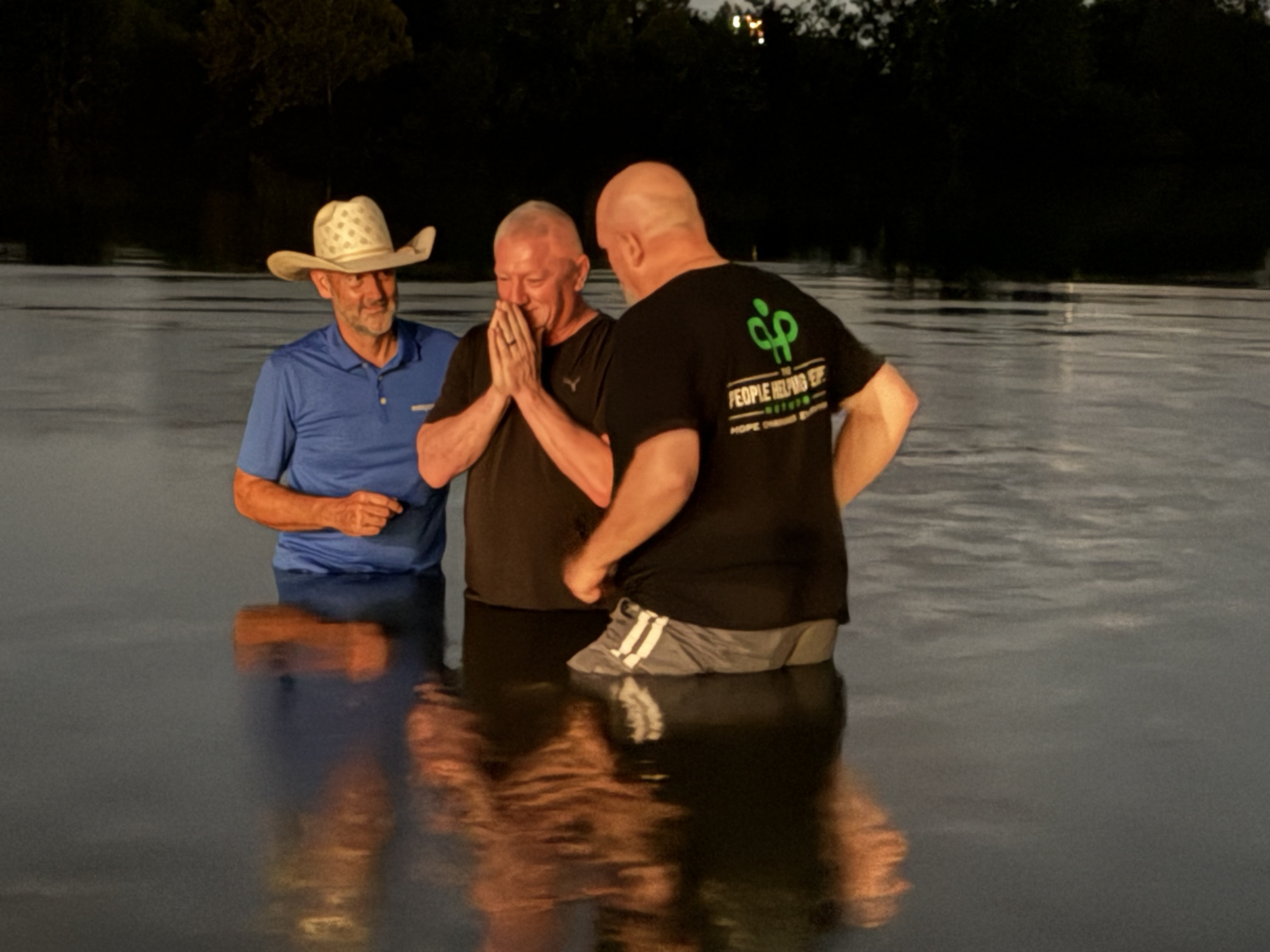 Three men, including one wearing a white cowboy hat, standing waist-deep in a lake during a sunset baptism