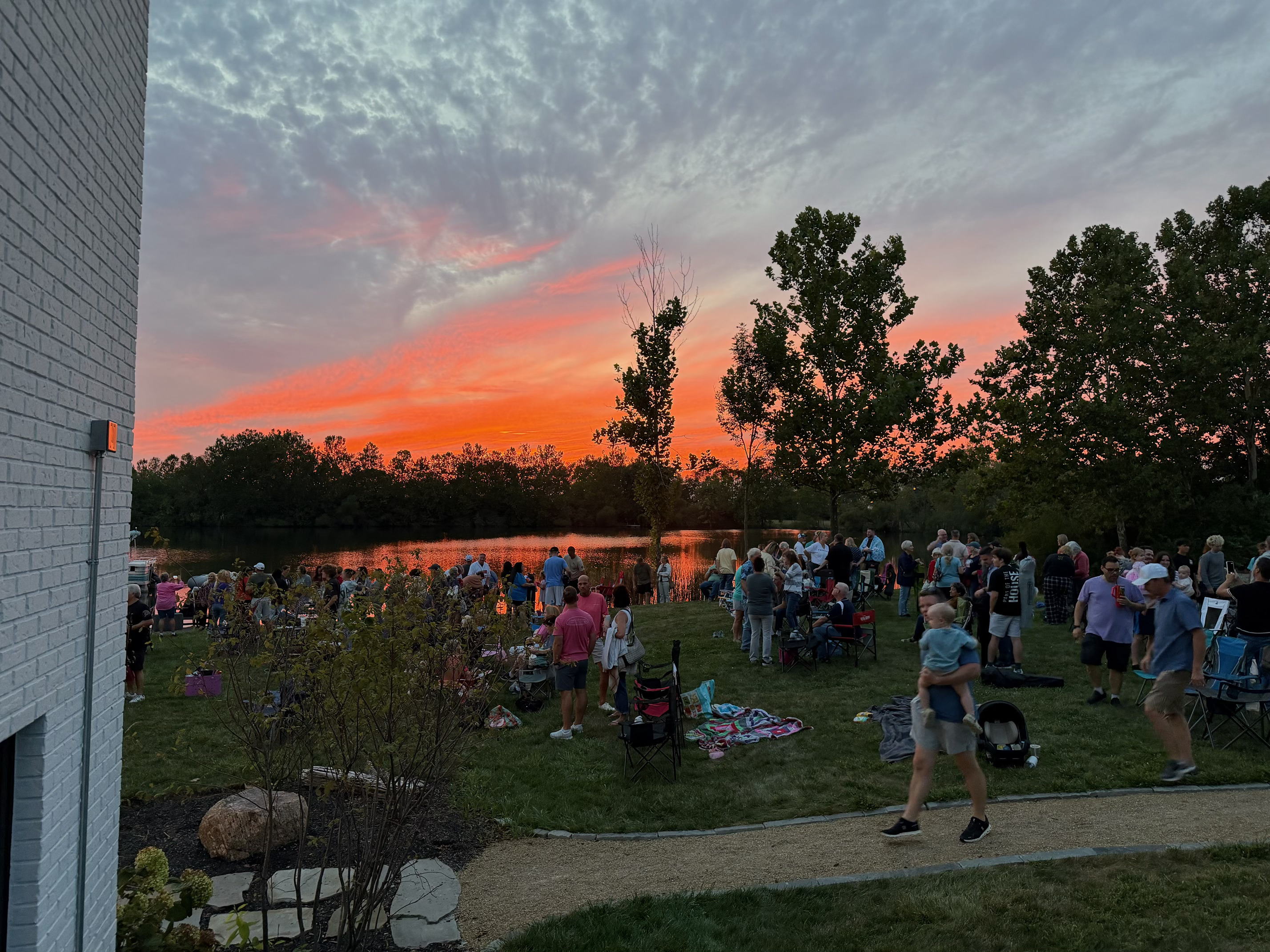 A large community gathering on a lawn by a lake, silhouetted against a brilliant orange and red sunset