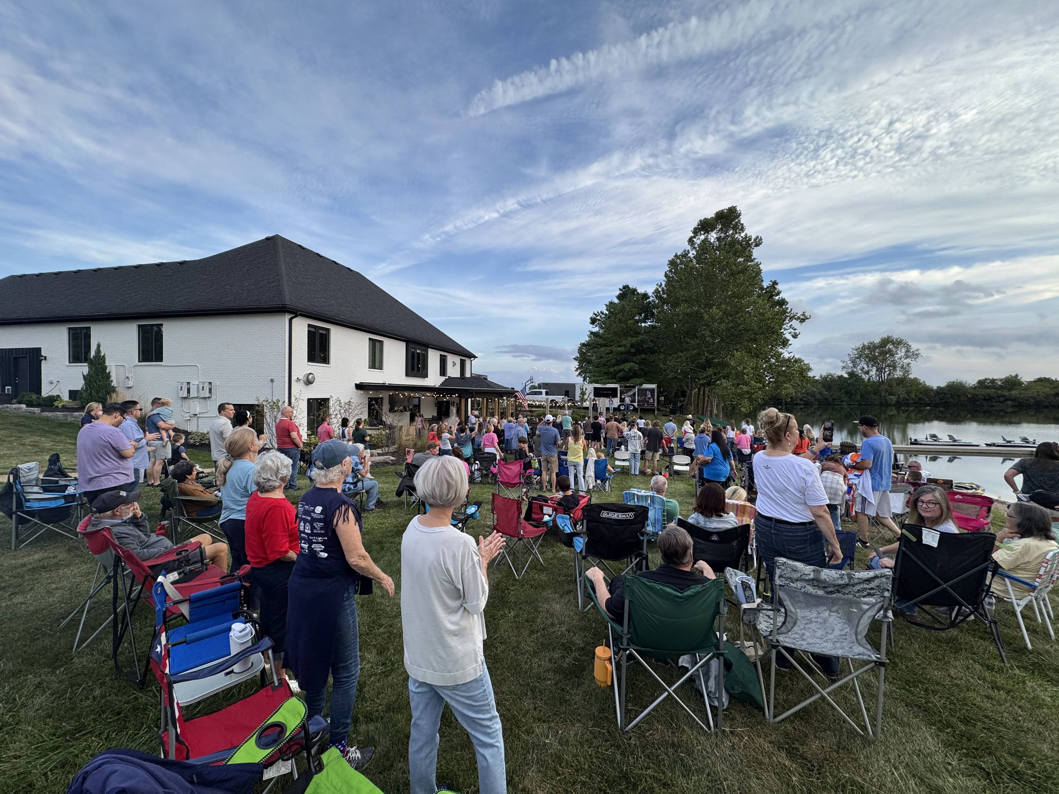 A congregation seated in lawn chairs for an outdoor service on a grassy slope between a white farmhouse and a lake