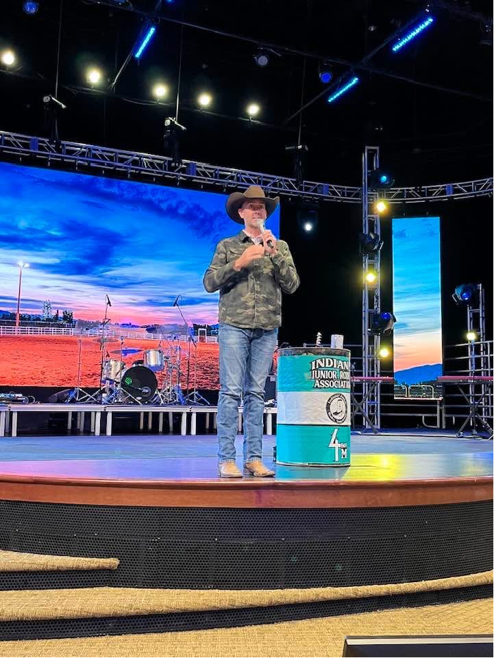 A speaker in a cowboy hat on a lit stage beside a rodeo barrel