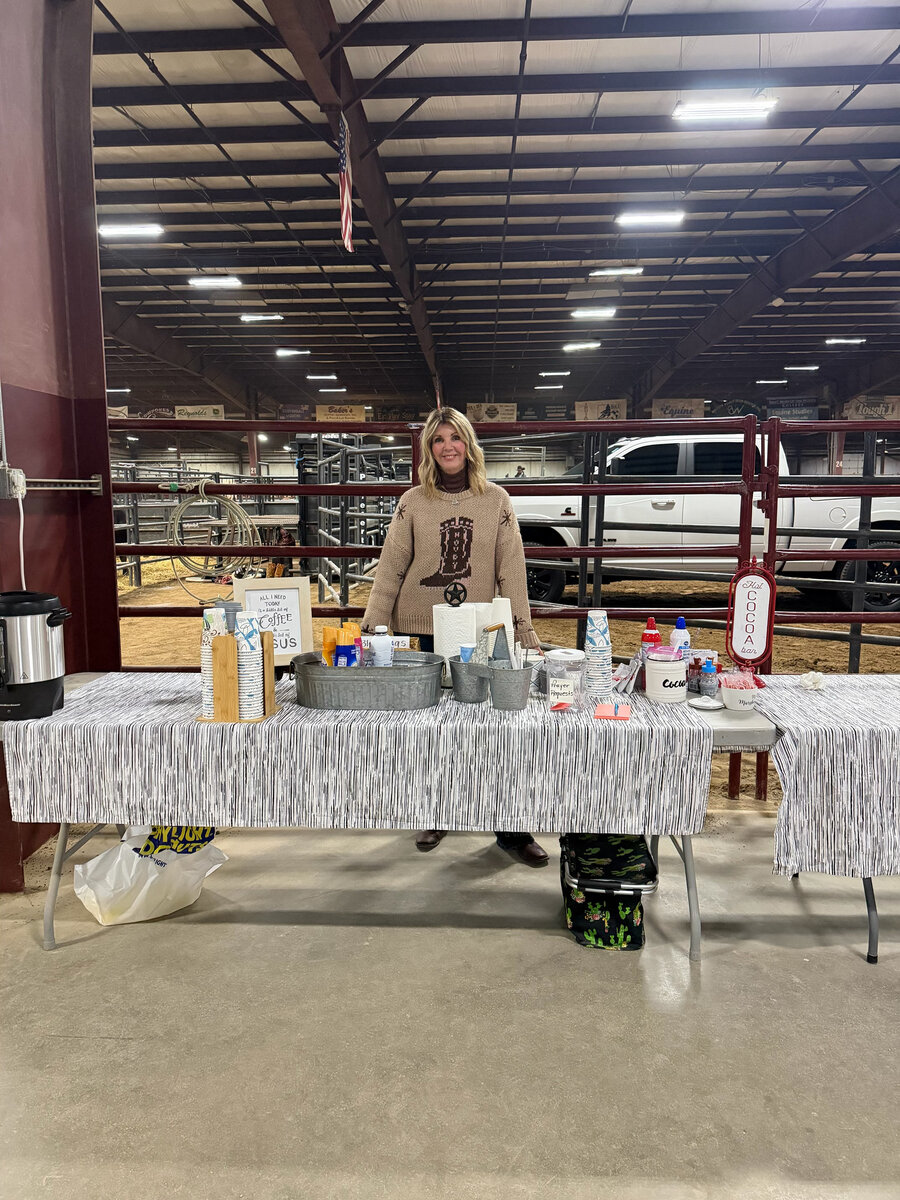 A woman standing behind a coffee and hot cocoa table set up at the arena