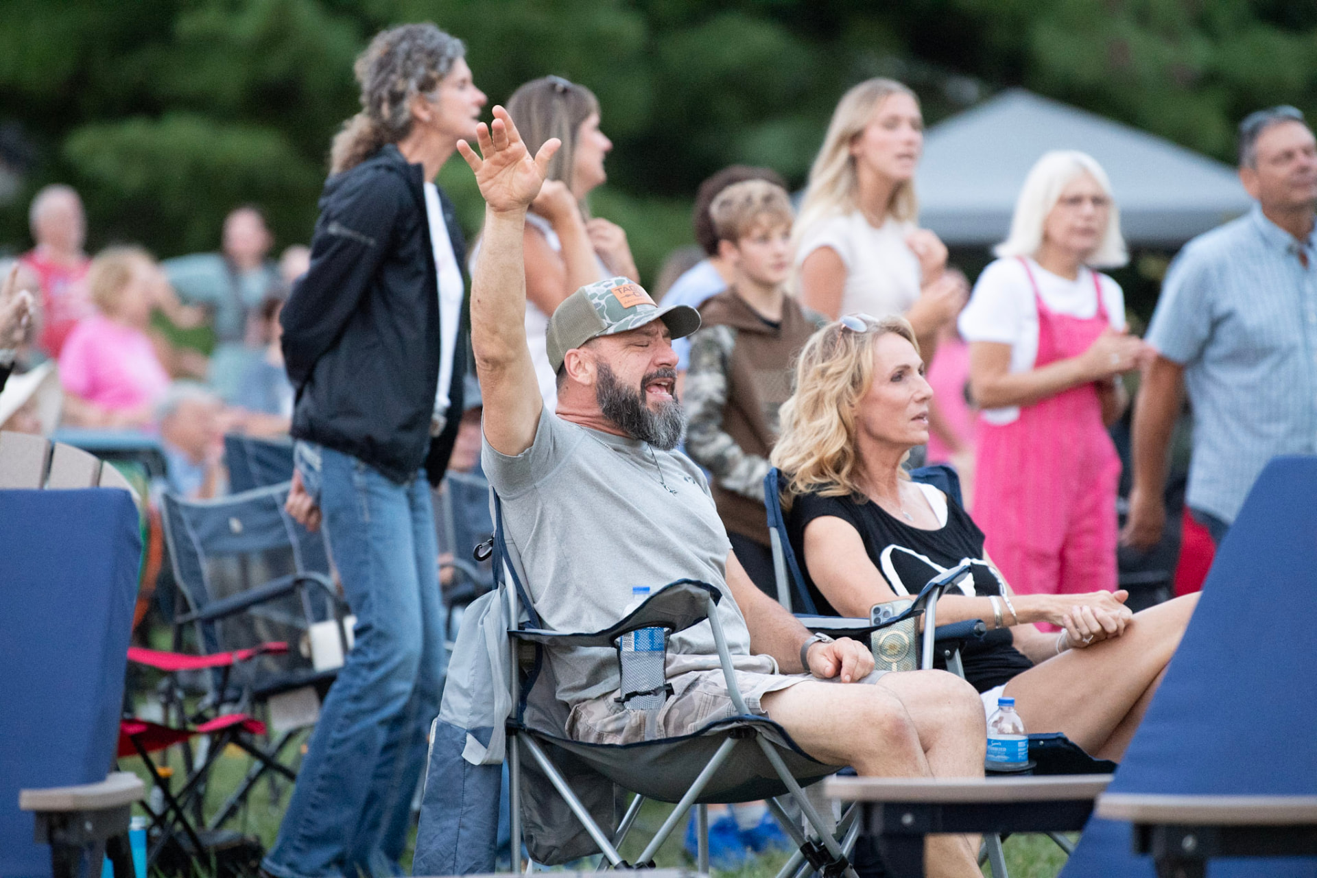 A man in a camouflage cap raising his arm in worship beside a crowd seated in folding chairs at an outdoor gathering