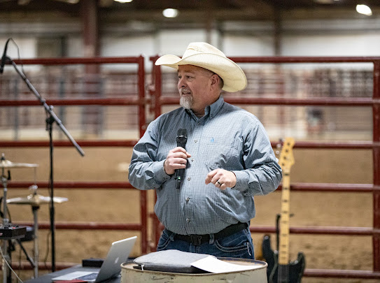 A man in a white cowboy hat speaking into a microphone beside a barrel pulpit